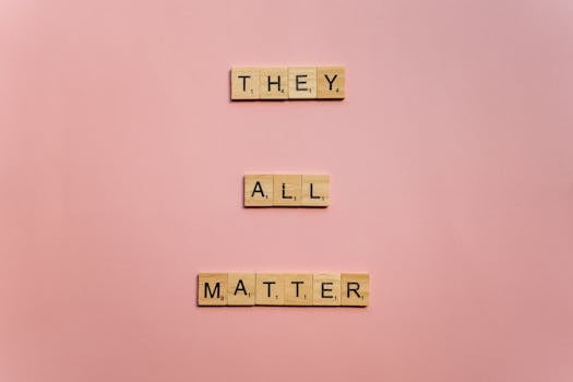 Wooden Scrabble tiles spell 'They All Matter' on a pink background, promoting inclusivity.
